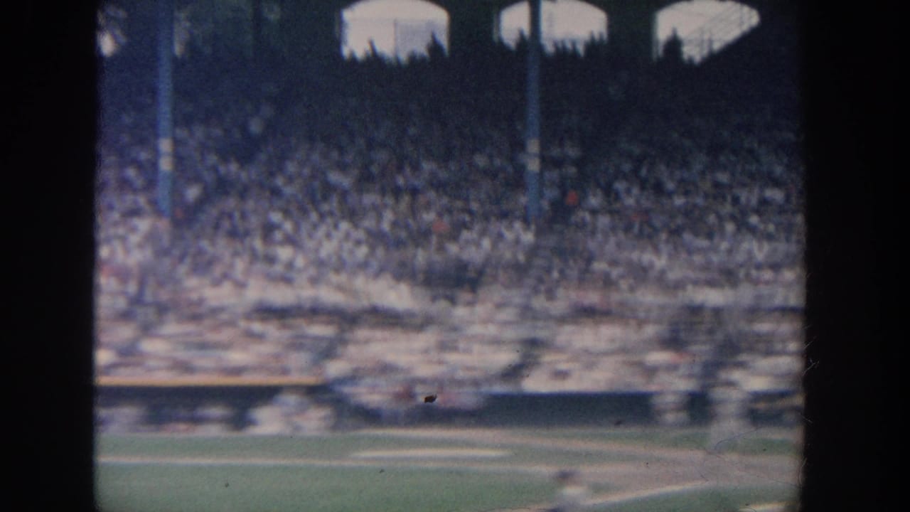 1964 New York baseball stadium view with a packed grandstand and blurred field action seen through dark framing.