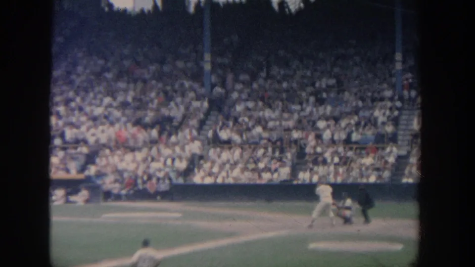 1964 New York 8mm frame showing home plate action with batter, catcher, umpire, fielder, and grandstand crowd.