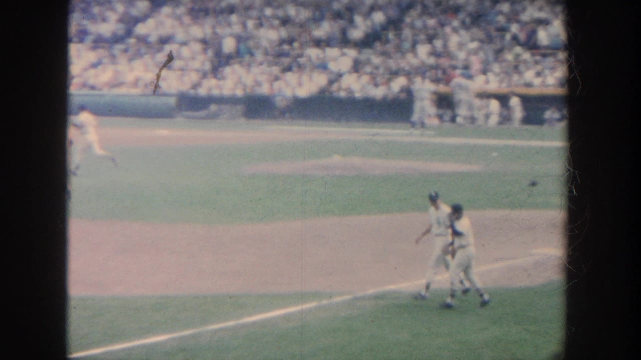 1964 New York baseball home-movie frame showing players in white uniforms near the infield and a dense crowd in the stands.
