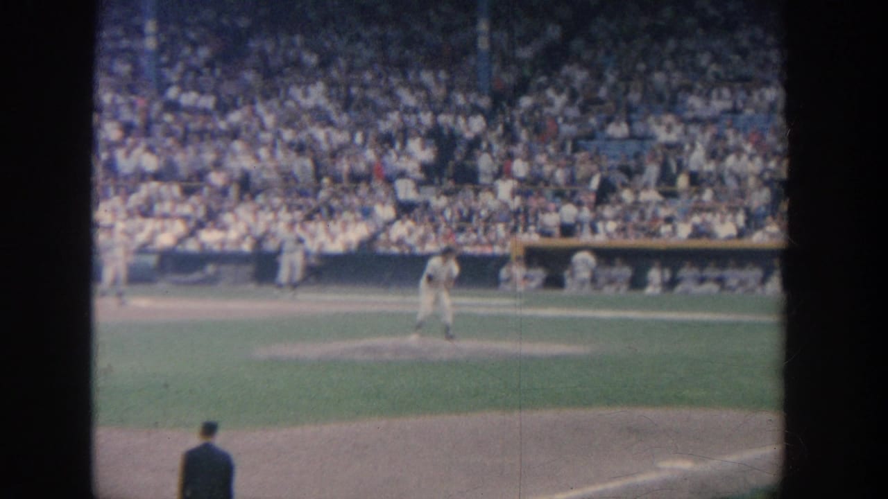 Pitcher on the mound with fielders and a large crowd visible in the stadium background.