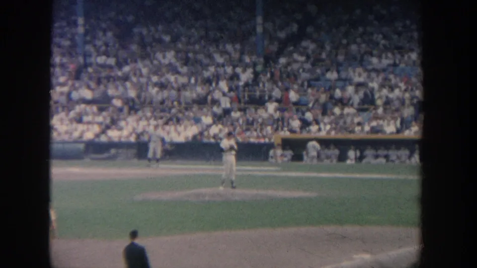 The frame shows a wide spectator view of a baseball game, with the mound, dugout benches, and crowded stands visible through 8mm film softness.