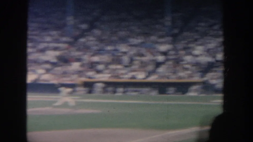 A soft, motion-blurred 8mm frame shows a baseball field edge, distant players, dugout benching, and packed stadium seating.