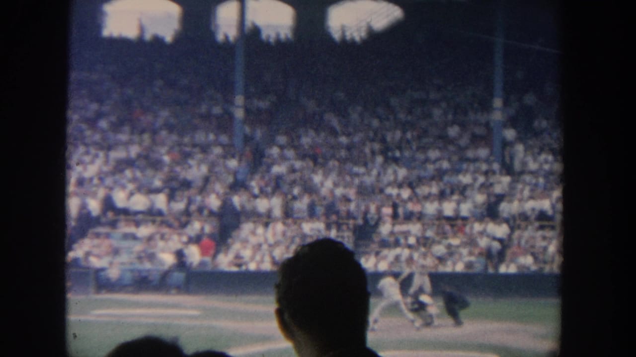 1964 New York baseball home-movie frame showing a batter near home plate, catcher, umpire, and packed stands beyond dark foreground framing.