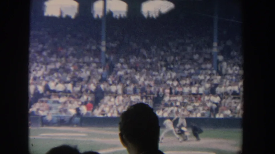 1964 New York baseball home-movie frame showing a batter near home plate, catcher, umpire, and packed stands beyond dark foreground framing.