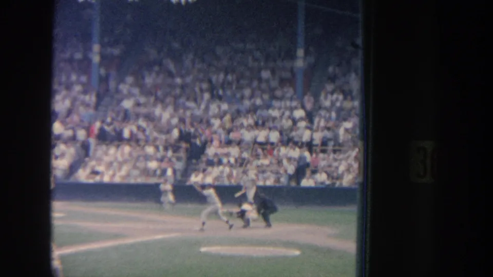 Grainy 1964 New York 8mm frame of baseball action at home plate, with the batter, catcher, umpire, and dense crowd visible.