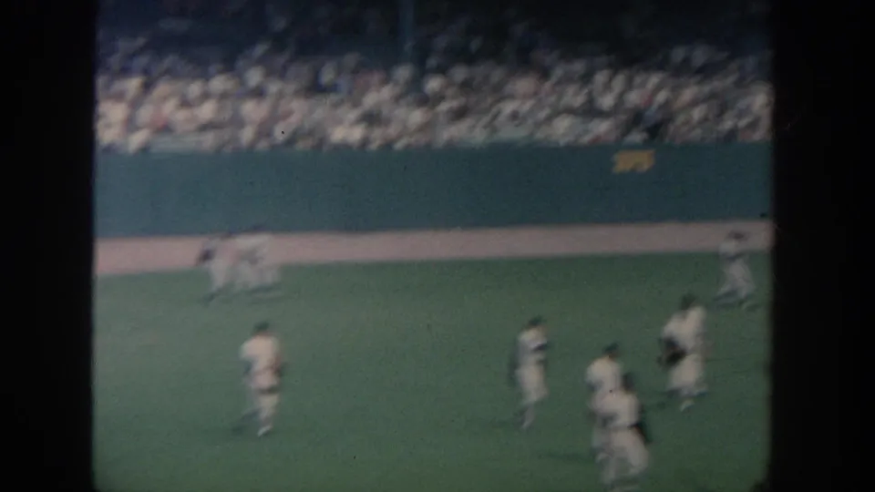 1964 New York baseball-stadium field view with players in white uniforms walking across the grass, a crowd in the stands, and a dark foreground obstruction crossing the frame.