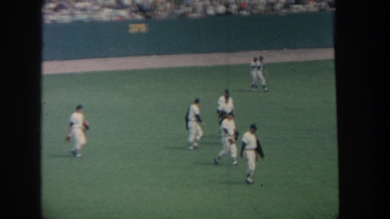 Baseball players in white uniforms walk across a green field near the outfield wall.