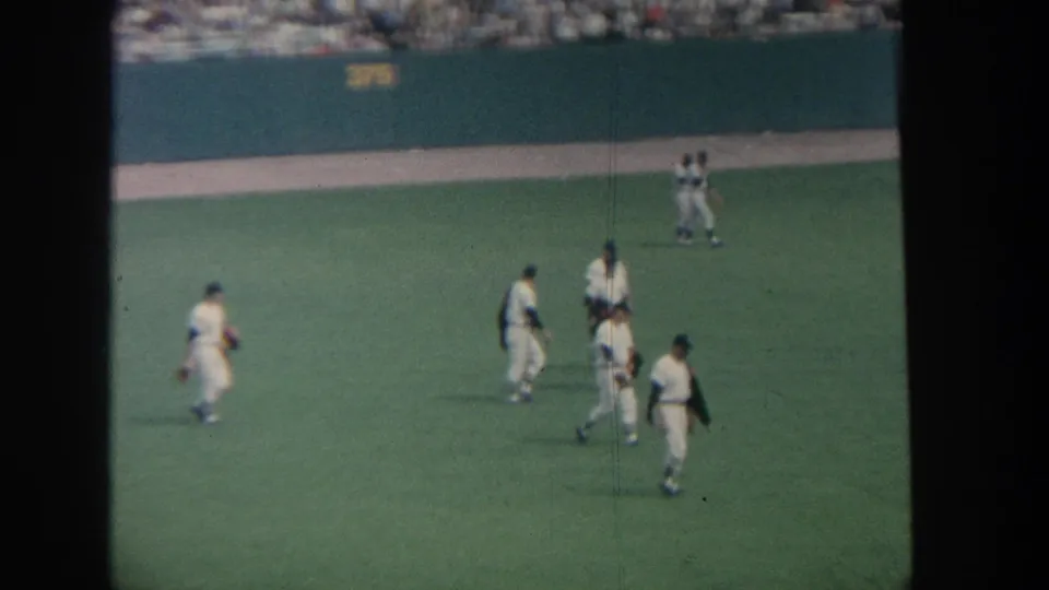 1964 New York home-movie view of baseball players in white uniforms crossing the field with spectators beyond the outfield wall.