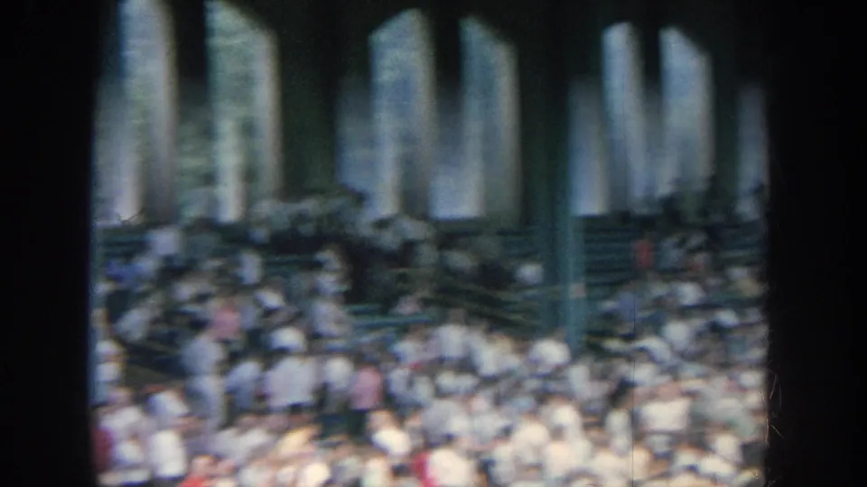Home-movie frame of spectators in stadium seating, partly obscured by vertical columns and soft motion blur.