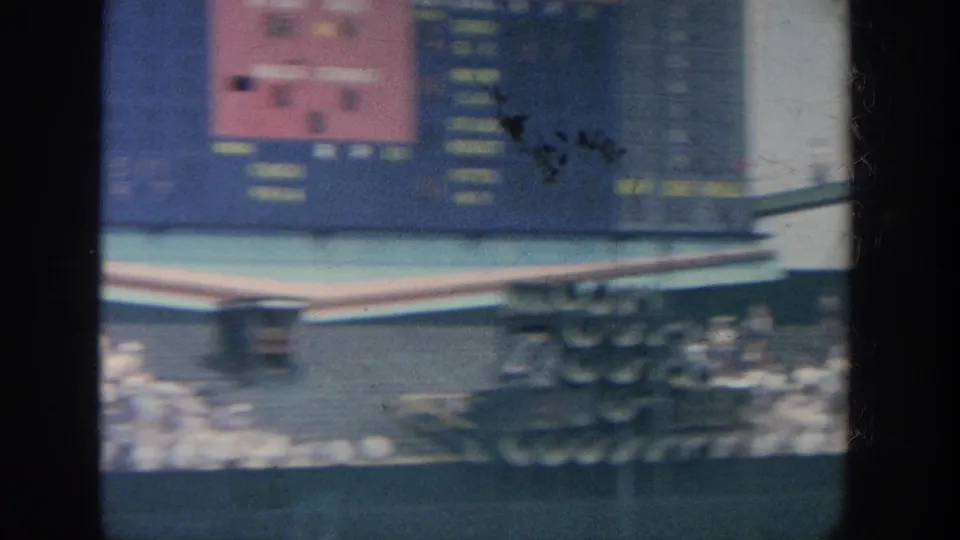 1964 New York home-movie frame showing a blurred ballpark scoreboard, packed stands, and a portion of the field from the spectator area.
