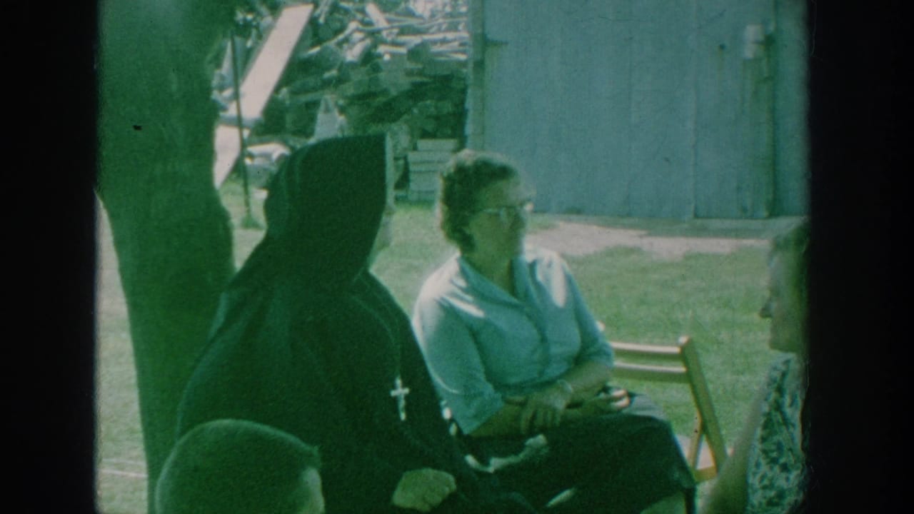 1964 New York home-movie frame showing a Catholic nun in a black habit with women seated on wooden chairs in a grassy yard.