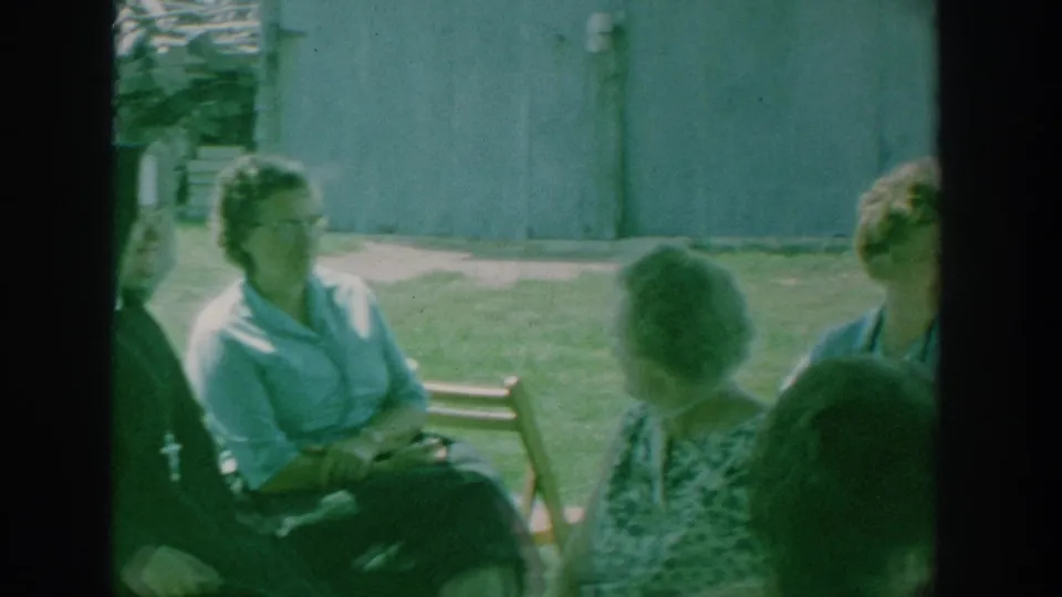 1964 New York footage frame with seated women facing one another outdoors, with a nun partly visible at the left side.