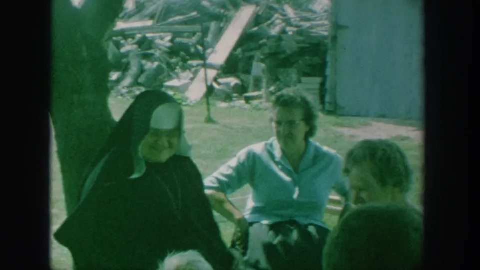 1964 New York home-movie frame showing a smiling nun and women seated close together outdoors near a shed.