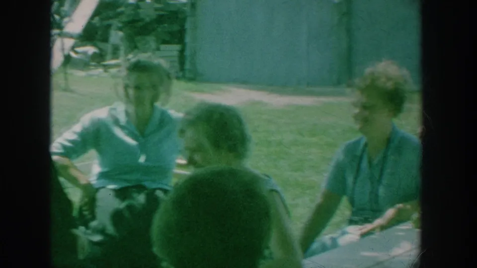 1964 New York home-movie frame showing women seated in conversation outdoors, with the nun mostly outside the left edge.