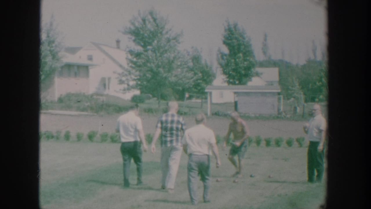 Several men stand on a lawn during a bocce game with houses and trees behind them.