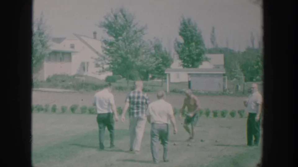 1964 New York 8mm frame of adult men on a suburban lawn during a bocce game, with houses, trees, and a planted row in the background.