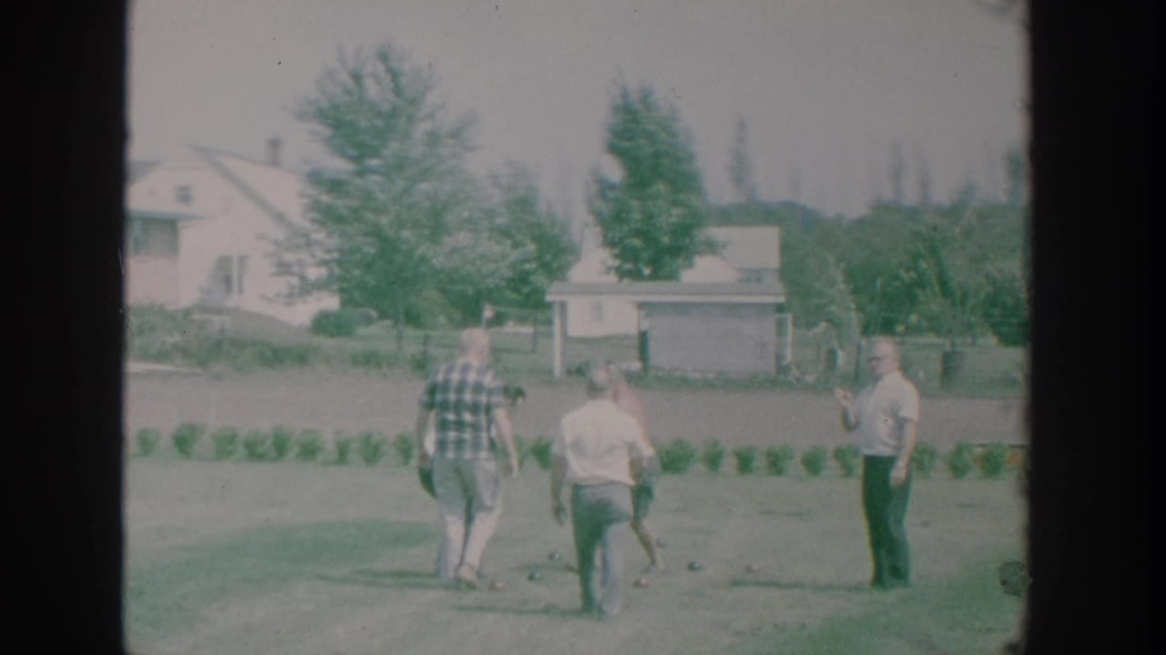 Adult men move around a suburban New York lawn during a 1964 bocce game, seen in soft 8mm color with nearby homes and trees.