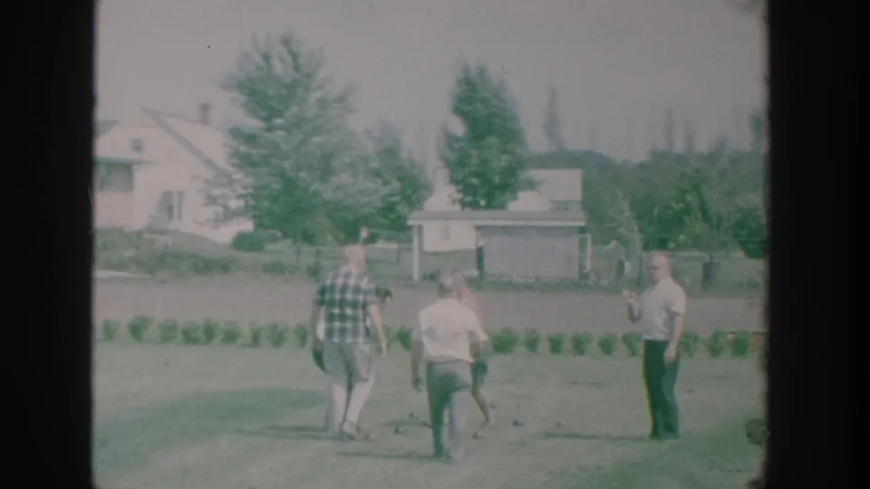 Adult men move around a suburban New York lawn during a 1964 bocce game, seen in soft 8mm color with nearby homes and trees.