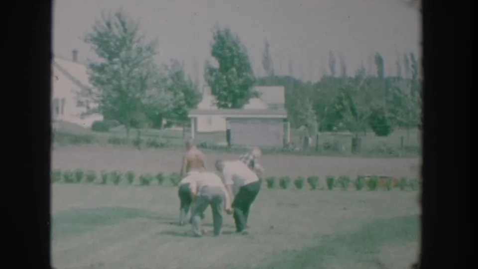 The bocce players bend together on a suburban lawn in a 1964 New York 8mm home movie, with residential houses and trees behind them.