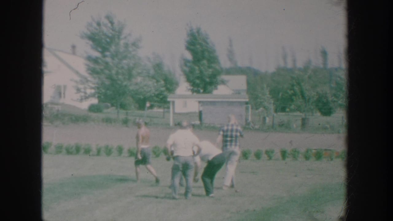 1964 New York home-movie frame showing older men playing bowls on a suburban backyard lawn with houses and trees beyond.