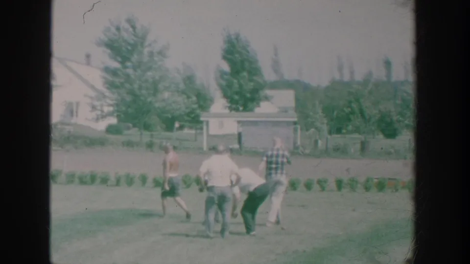 1964 New York home-movie frame showing older men playing bowls on a suburban backyard lawn with houses and trees beyond.