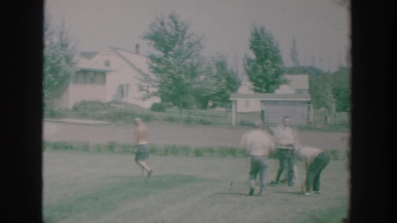 Men in casual clothes watch and move across a backyard lawn during a bowls game.