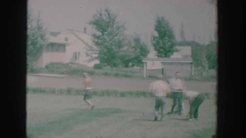 Older men in summer clothing continue a bowls game on a grass lawn, with a low house and trees in the background.