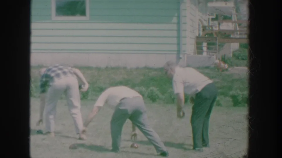 Close backyard view of older men measuring or picking up bowls near a row of plants and a green-sided house.