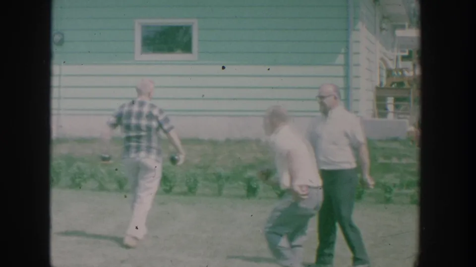 End frame of the 1964 backyard bowls scene, with older men walking on the lawn beside a green-sided suburban house.