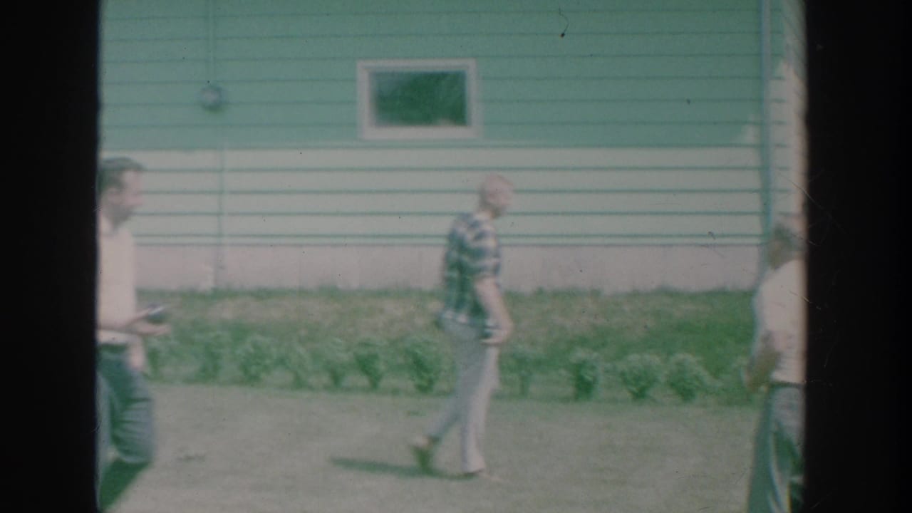 1964 New York 8mm frame showing men walking past a green-and-white clapboard house in a yard.