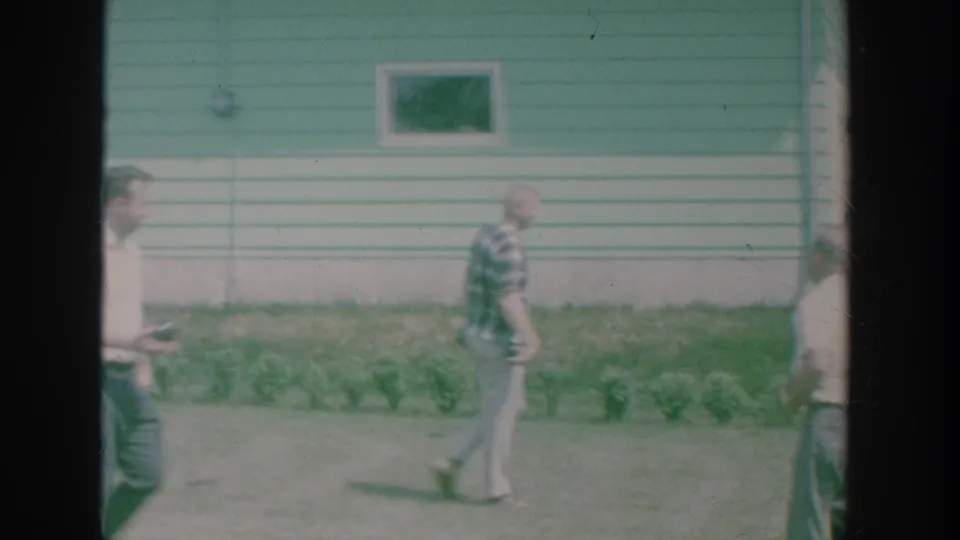 1964 New York 8mm frame showing men walking past a green-and-white clapboard house in a yard.