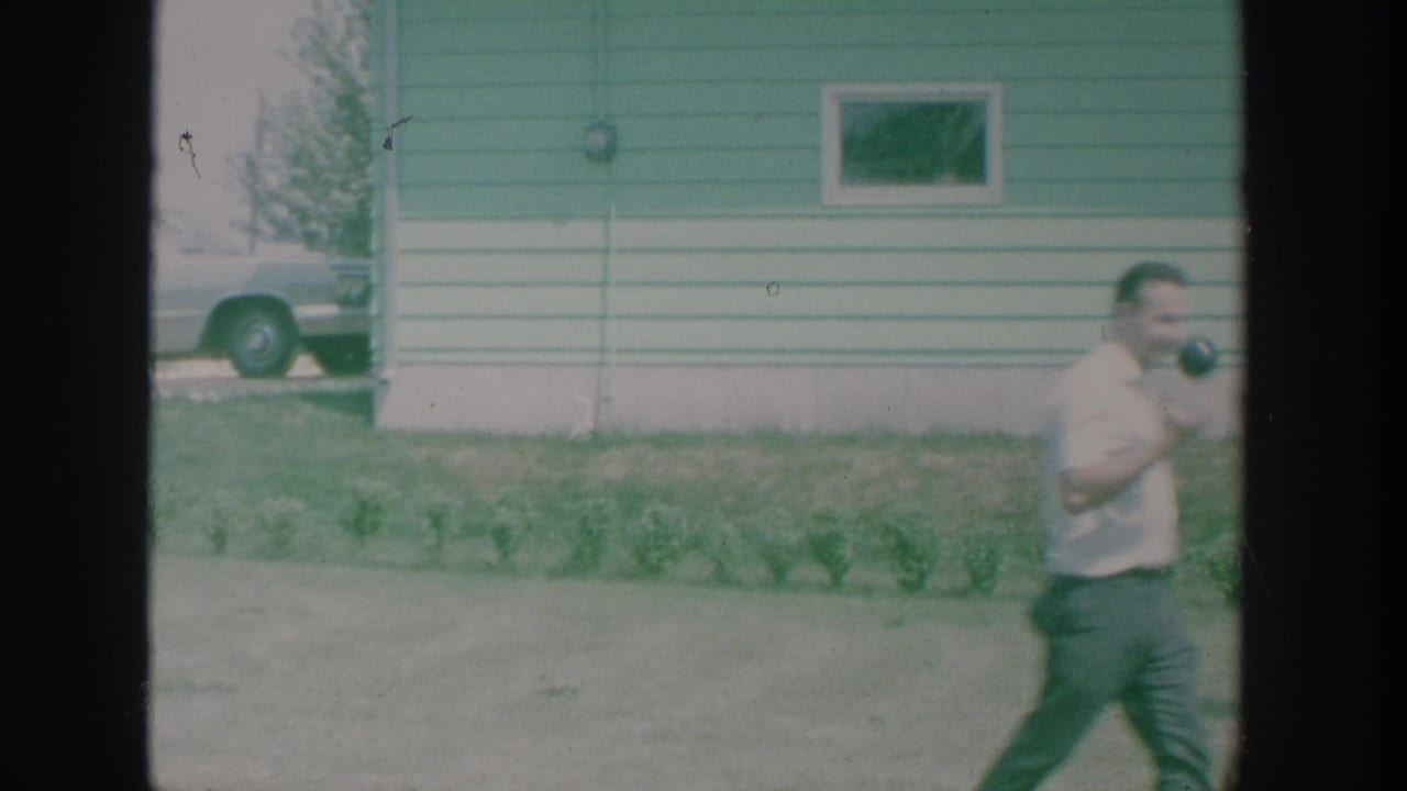 Man carrying a camera walks past the side of a house with a parked car nearby.
