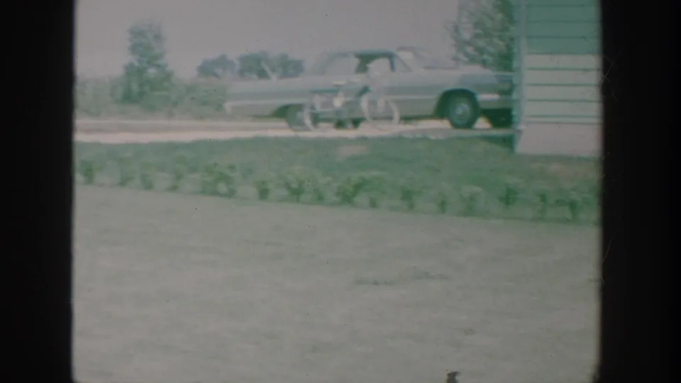 A parked mid-century sedan and bicycle appear near the corner of a green-and-white house in an 8mm yard scene.