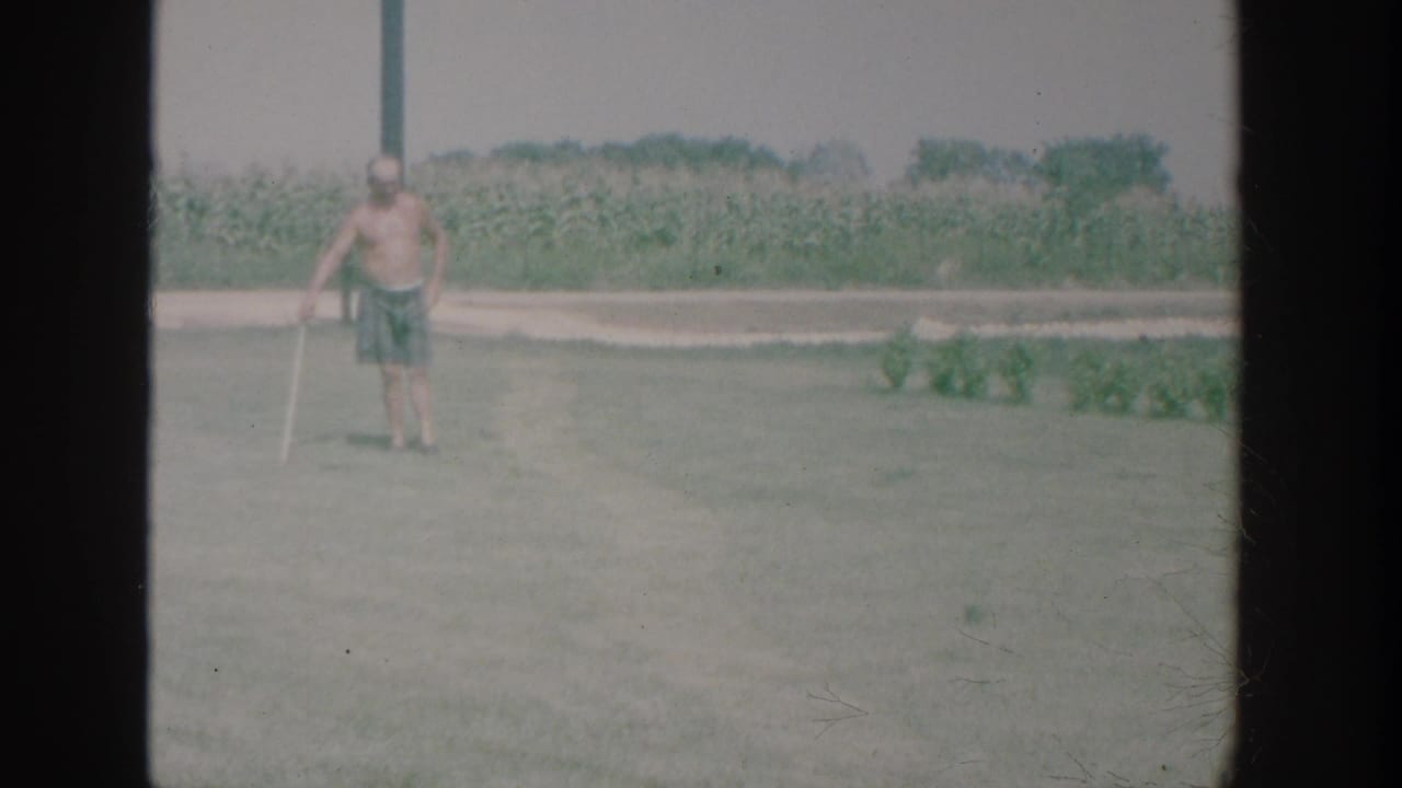 Shirtless man holding a croquet mallet on a lawn with a cornfield behind him.
