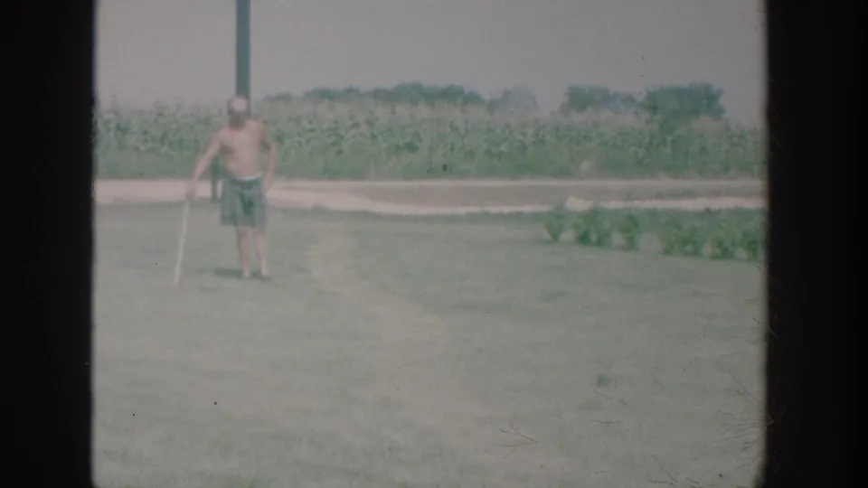 1964 New York home-movie frame showing a shirtless man in swim trunks standing with a croquet mallet on a grassy lawn near a cornfield.