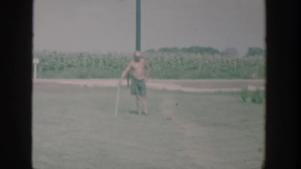 1964 New York home-movie frame of a shirtless man playing croquet on a broad lawn with a cornfield and road edge behind him.