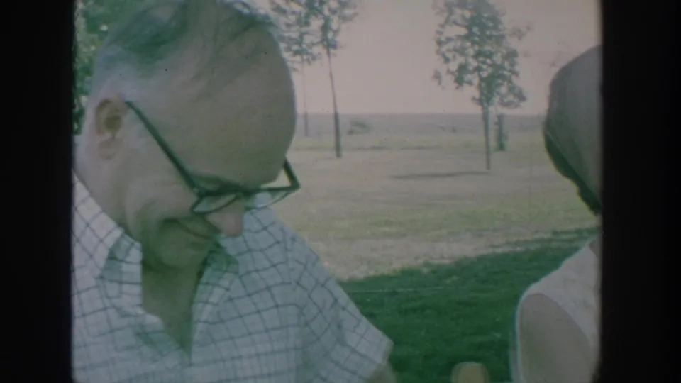1964 New York home-movie frame showing a smiling man in glasses and a checkered shirt with other adults nearby on a lawn.