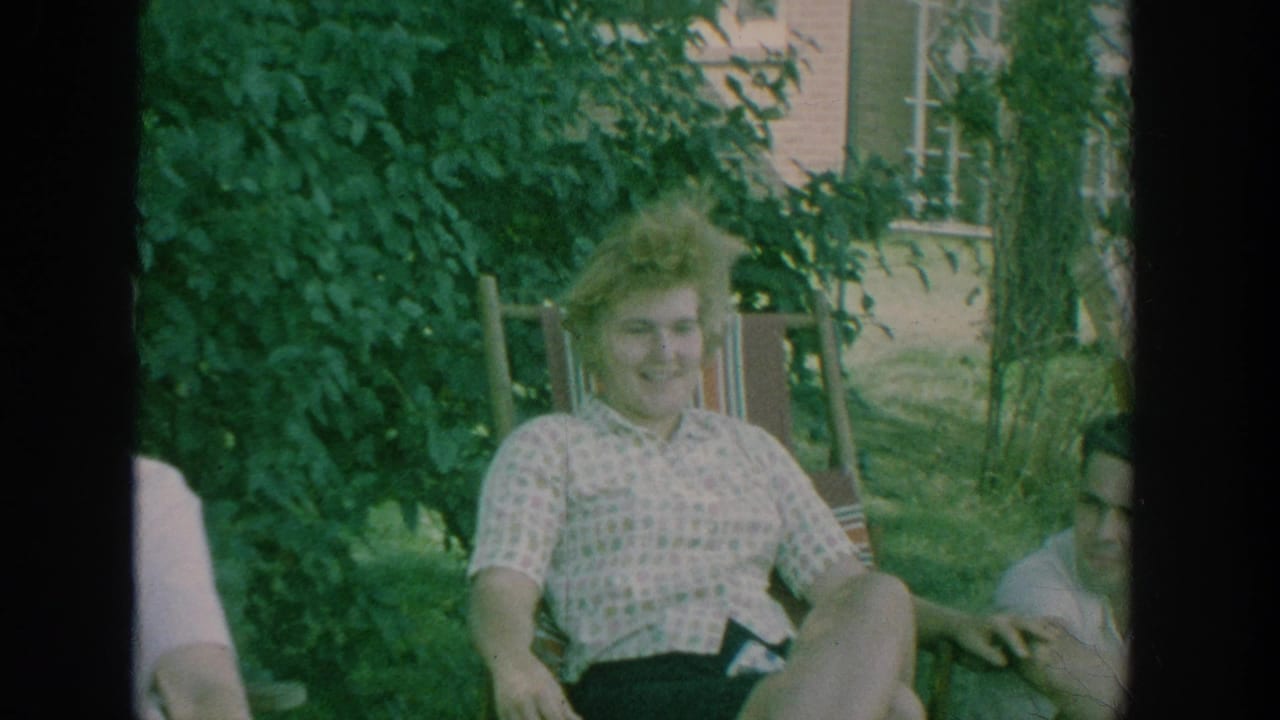 1964 New York home-movie frame showing adults relaxing in a shaded garden beside a brick house, with a woman smiling toward the camera.