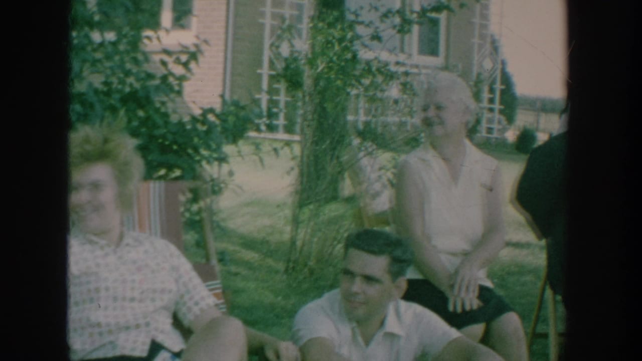 1964 New York home-movie frame showing adults gathered on a lawn, with garden plants and house windows behind them.