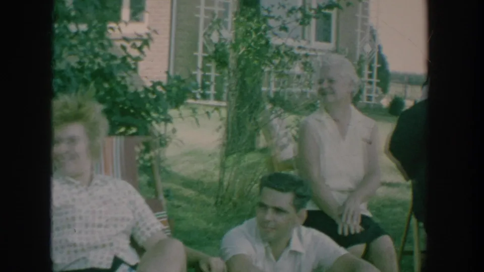 1964 New York home-movie frame showing adults gathered on a lawn, with garden plants and house windows behind them.