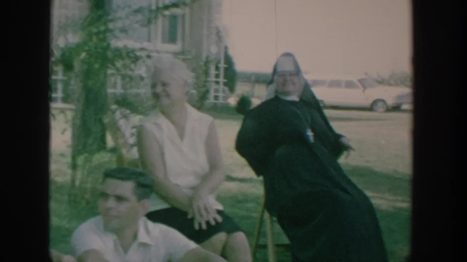 1964 New York home-movie frame showing a nun seated outdoors with adults during a relaxed garden gathering.