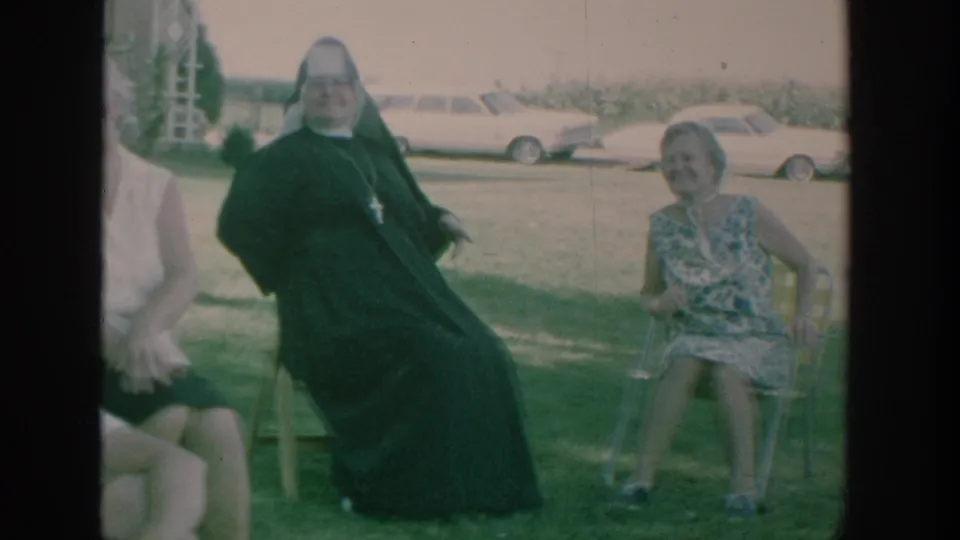 1964 New York home-movie frame showing a nun and a woman seated on lawn chairs, with older parked cars behind them.