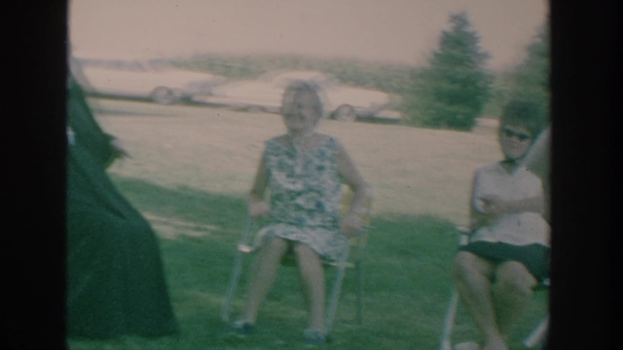 Adults seated on folding chairs in a grassy yard, with older cars in the background.