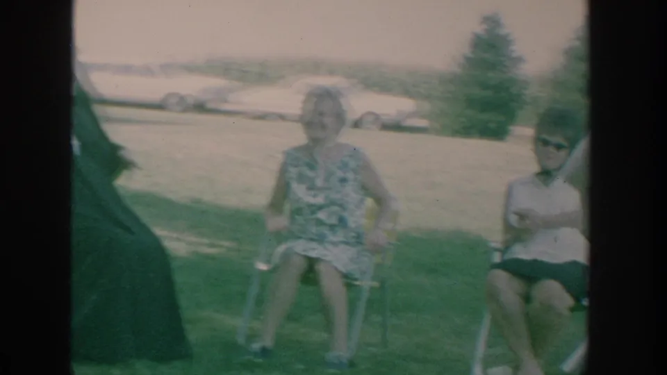 1964 New York 8mm frame showing adults seated on lawn chairs during an outdoor family or friend gathering.