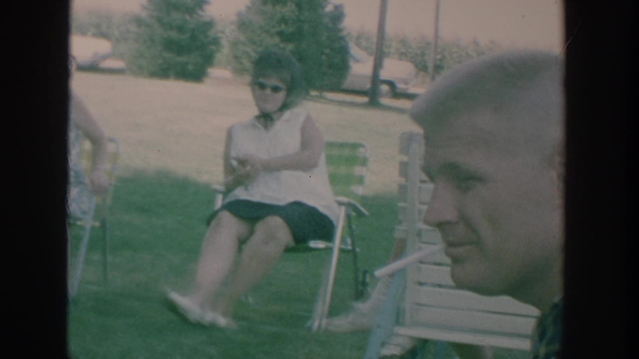 1964 New York home-movie frame with a man smoking in the foreground and adults seated outdoors behind him.