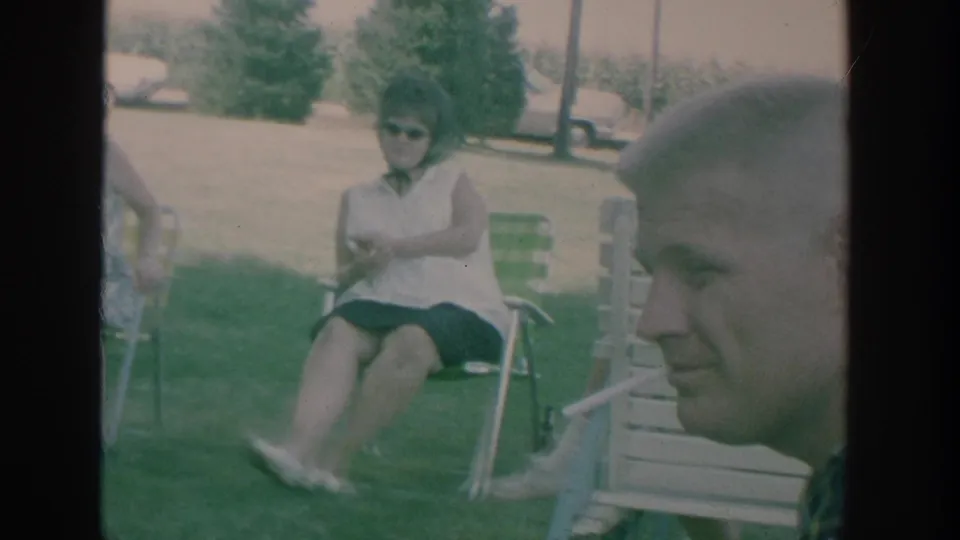 1964 New York home-movie frame with a man smoking in the foreground and adults seated outdoors behind him.
