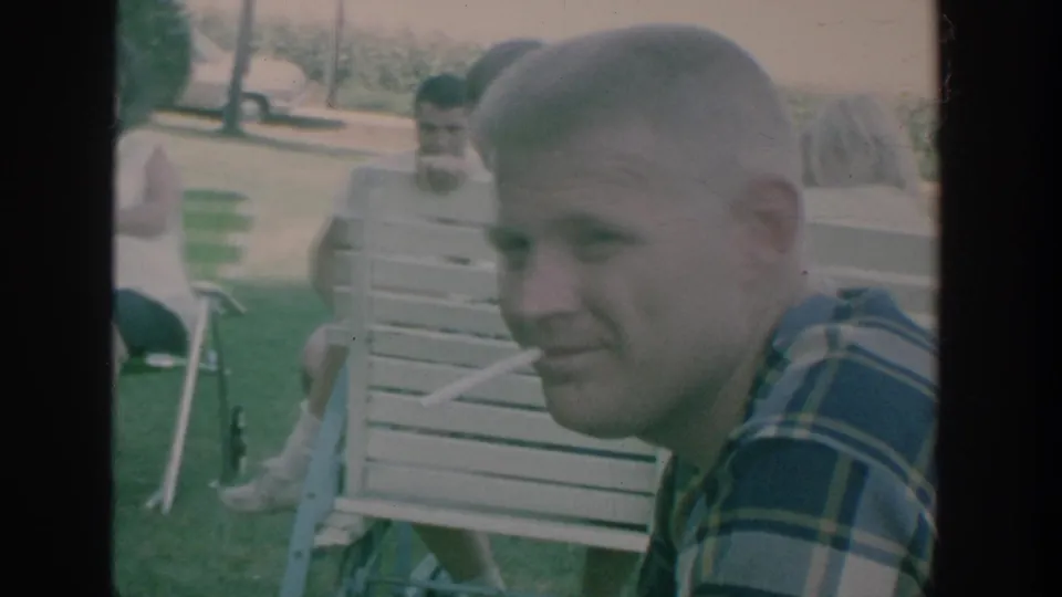 1964 New York 8mm frame showing a man in a plaid shirt near folding chairs during an outdoor gathering.
