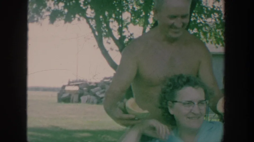 1964 New York home-movie frame with a shirtless man holding watermelon near a smiling woman with glasses in a yard.