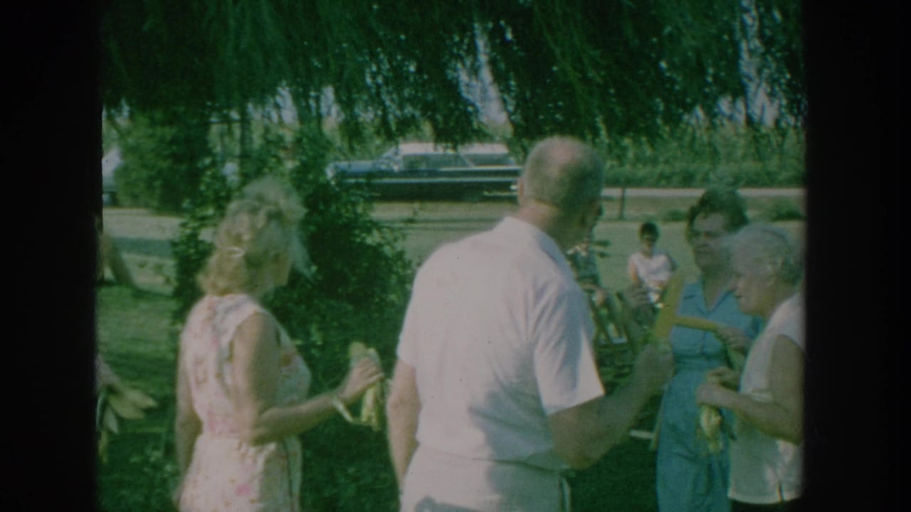 1964 New York home-movie frame showing adults under a large tree, a woman in a floral dress, and corn being handled during an outdoor gathering.