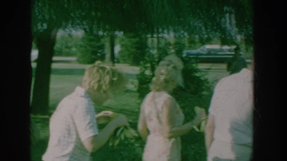 1964 New York footage frame with adults in summer clothing near a bench or chair, partly obscured by tree shade and film exposure.
